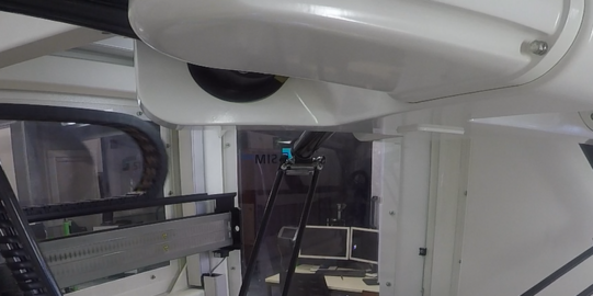Foto einer industriellen Maschine in einem Laborumfeld. Die Maschine besteht aus mehreren Roboterarmen, die an einem zentralen Punkt zusammenlaufen und auf einem Arbeitstisch operieren. Der Roboter ist in der Mitte des Bildes fokussiert und führt präzise Bewegungen aus, um auf kleinen blauen Platten zu arbeiten. Im Hintergrund sind verschiedene technische Geräte und Computer zu sehen, die die komplexe Arbeitsumgebung ergänzen.
