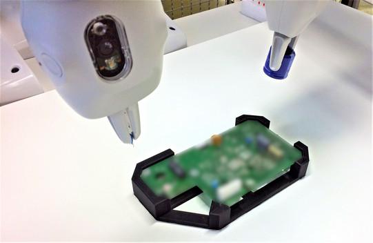 Foto von zwei Roboterarmen, die über einer unscharfen elektronischen Platine arbeiten. Die Roboterarme sind weiß und haben präzise Werkzeuge an ihren Enden. Einer der Arme hat ein blaues Werkzeug, während der andere ein weißes Werkzeug mit einer Kamera oder einem Sensor besitzt. Die Platine liegt in einer schwarzen Halterung auf einer weißen Arbeitsfläche. Die Szene deutet auf einen automatisierten Montage- oder Inspektionsprozess in einer hochmodernen Produktionsumgebung hin.
