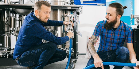 Das Bild zeigt zwei Männer, die vor einer industriellen Anlage arbeiten. Beide tragen blaue, karierte Hemden und sind in der Hocke. Der Mann links hat kurze, braune Haare und hält ein Ventil an der Anlage. Der Mann rechts hat längeres Haar und einen Bart, er hält einen blauen Schlauch. Im Hintergrund sind weitere metallische Maschinen und Geräte zu sehen. Der Arbeitsplatz wirkt technisch und industriell.