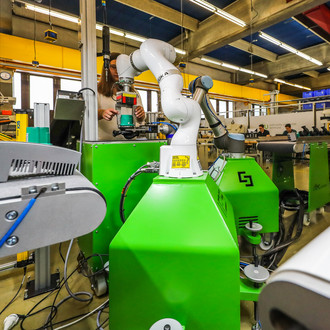 Foto: Eine industrielle Werkstatt mit verschiedenen grünen Maschinen und einem weißen Roboterarm in der Mitte. Der Roboterarm führt präzise Arbeiten an einem der Maschinen durch. Die Umgebung ist hell beleuchtet und modern ausgestattet, mit mehreren technischen Geräten und Kabeln. Im Hintergrund sind weitere Arbeitsbereiche und Personen zu erkennen, die an anderen Maschinen arbeiten. Die Decke ist mit farbigen Schallschutzpaneelen versehen.