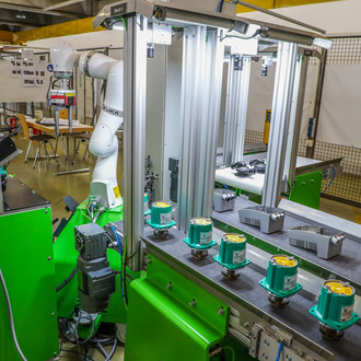 Foto einer industriellen Roboterstation. Im Vordergrund sind grüne Maschinen mit mehreren Bauteilen auf einem Förderband zu sehen. Ein Roboterarm befindet sich in der Mitte des Bildes und ist von Metallgestellen umgeben. Im Hintergrund sind weitere industrielle Ausrüstungen sowie Informationstafeln und Stühle zu erkennen.