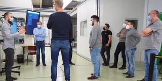 Foto einer Gruppe von Männern in einer industriellen Umgebung, die sich um einen Bildschirm versammelt haben. Der Bildschirm zeigt eine Präsentation oder Schulung. Alle Personen tragen Gesichtsmasken und stehen in einem Abstand zueinander, was auf COVID-19-Schutzmaßnahmen hinweist. Der Mann links vorne gestikuliert und scheint etwas zu erklären, während die anderen aufmerksam zuhören. Im Hintergrund sind industrielle Geräte und Kabinen zu sehen, die die technische Umgebung verdeutlichen. Die Männer tragen überwiegend Freizeitkleidung und wirken konzentriert und interessiert.