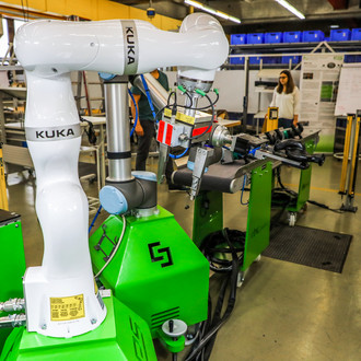 Industriehalle mit einem großen, weißen KUKA-Roboterarm, der auf einer grünen Plattform montiert ist. Der Roboterarm führt eine Aufgabe aus und hält ein Werkzeug. Im Hintergrund sind weitere Maschinen, Arbeitsstationen und eine Person zu sehen, die an einem Tisch arbeitet. Der Raum ist hell beleuchtet und gut organisiert, mit verschiedenen Produktionsgeräten und Arbeitsmaterialien.