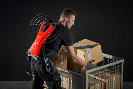 Foto eines Mannes, der ein Exoskelett trägt und eine Kiste anhebt. Der Mann, mit schwarzem T-Shirt und Handschuhen bekleidet, beugt sich vor, um eine Kiste aus einem Gitterwagen zu heben. Das Exoskelett ist rot und schwarz und unterstützt den Rücken des Mannes, während er die Kiste anhebt.
