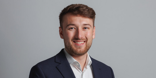 Foto eines jungen Mannes vor einem Hintergrund mit den Buchstaben "IPS". Der Mann hat kurzes, braunes Haar und trägt ein weißes Hemd sowie ein graues Sakko. Er lächelt freundlich in die Kamera. Der Hintergrund ist in hellen Farben gehalten, wobei die Buchstaben "IPS" in einem großen, grünen Schriftzug zu sehen sind.