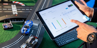 Das Bild zeigt eine Person, die ein Tablet in den Händen hält und auf dem Bildschirm Diagramme und Daten betrachtet. Im Hintergrund ist eine Modellautorennbahn mit zwei Spielzeugautos zu sehen, einem blauen und einem weißen. Die Bahn hat Kurven und eine Ziellinie, und es sind weitere Elemente wie ein Steuergerät sichtbar. Die Person trägt eine Uhr am linken Handgelenk und scheint sich auf die Daten auf dem Tablet zu konzentrieren.