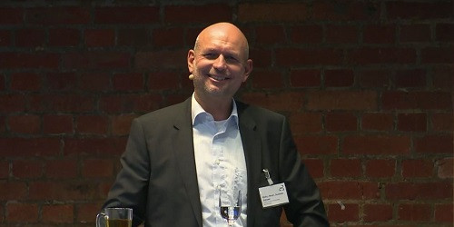 Foto eines lächelnden Mannes, der vor einer roten Ziegelwand steht. Der Mann hat eine Glatze und trägt einen schwarzen Anzug mit einem weißen Hemd. Er hat ein Namensschild an der linken Seite seines Anzugs und ein Mikrofon am Kragen befestigt. Vor ihm auf einem Tisch stehen ein Glas Wasser und ein Glas Tee. Der Mann wirkt entspannt und freundlich, was darauf hindeutet, dass er möglicherweise einen Vortrag oder eine Präsentation hält.