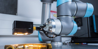 Das Bild zeigt einen modernen Industrieroboterarm, der ein goldfarbenes, metallisches Objekt inspiziert. Der Roboterarm, der blau-graue Akzente aufweist, ist mit einer Kamera oder einem Sensor ausgestattet, der das Objekt beleuchtet und untersucht. Der Hintergrund deutet auf eine industrielle oder Laborumgebung hin, mit verschiedenen technischen Geräten und einer Arbeitsfläche. Das Bild vermittelt einen Eindruck von Präzision und fortschrittlicher Technologie in der industriellen Fertigung oder Qualitätskontrolle.