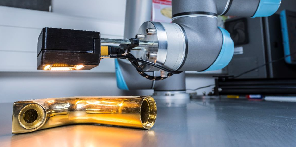 Das Bild zeigt einen modernen Industrieroboterarm, der ein goldfarbenes, metallisches Objekt inspiziert. Der Roboterarm, der blau-graue Akzente aufweist, ist mit einer Kamera oder einem Sensor ausgestattet, der das Objekt beleuchtet und untersucht. Der Hintergrund deutet auf eine industrielle oder Laborumgebung hin, mit verschiedenen technischen Geräten und einer Arbeitsfläche. Das Bild vermittelt einen Eindruck von Präzision und fortschrittlicher Technologie in der industriellen Fertigung oder Qualitätskontrolle.