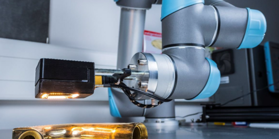 Das Bild zeigt einen modernen Industrieroboterarm, der ein goldfarbenes, metallisches Objekt inspiziert. Der Roboterarm, der blau-graue Akzente aufweist, ist mit einer Kamera oder einem Sensor ausgestattet, der das Objekt beleuchtet und untersucht. Der Hintergrund deutet auf eine industrielle oder Laborumgebung hin, mit verschiedenen technischen Geräten und einer Arbeitsfläche. Das Bild vermittelt einen Eindruck von Präzision und fortschrittlicher Technologie in der industriellen Fertigung oder Qualitätskontrolle.