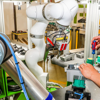 Foto von zwei Roboterarmen in einer industriellen Produktionslinie. Der weiße Roboterarm von KUKA ist mit einem Greifwerkzeug ausgestattet und hält ein Bauteil. Der graue Roboterarm mit blauen Kabeln befindet sich links im Bild. Rechts im Bild sind die Hände eines Arbeiters zu sehen, der mit einem roten Schraubenzieher an einem grünen Bauteil arbeitet. Die Umgebung ist eine moderne Werkstatt oder Fertigungsstätte mit verschiedenen Werkzeugen und Materialien im Hintergrund. Die Szene zeigt die Zusammenarbeit von Mensch und Maschine bei der Montage von Bauteilen.
