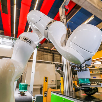Foto eines KUKA-Industrieroboters in einer Produktionsumgebung. Der weiße Roboterarm hat mehrere Gelenke und endet in einem Greifer, der ein Bauteil hält. Der Hintergrund zeigt eine industrielle Halle mit verschiedenen Maschinen und Regalen. Die Decke ist mit roten und blauen Paneelen verkleidet. Der Roboterarm ist auf eine grüne Arbeitsfläche ausgerichtet, die Teil einer größeren Produktionsanlage zu sein scheint. Die gesamte Szene vermittelt einen Eindruck von moderner Automatisierung und fortschrittlicher Fertigungstechnik.