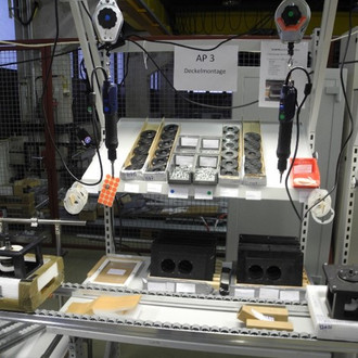 Das Bild zeigt einen Arbeitsplatz in einer industriellen Umgebung, der für die Montage eingerichtet ist. Über dem Arbeitstisch hängen zwei Elektrowerkzeuge an Halterungen. Auf dem Tisch befinden sich mehrere Bauteile und Werkzeuge, ordentlich in Reihen angeordnet. Ein Schild über dem Tisch trägt die Aufschrift "AP 3 Deckelmontage". Im Hintergrund sind weitere industrielle Geräte und eine Gitterabtrennung zu sehen.