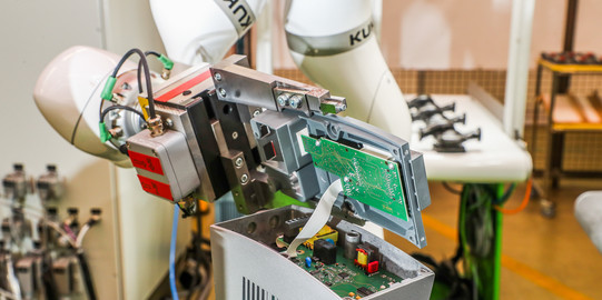 Das Bild zeigt eine Nahaufnahme eines industriellen Roboters, der an einer elektronischen Baugruppe arbeitet. Der Roboterarm ist weiß und trägt das Logo des Herstellers KUKA. Der Roboter hält eine teilweise geöffnete Baugruppe, die grüne Leiterplatten und verschiedene elektronische Komponenten enthält. Kabel und mechanische Teile sind ebenfalls sichtbar. Im Hintergrund sind weitere industrielle Ausrüstungen und Arbeitsbereiche zu sehen. Der Arbeitsplatz ist gut organisiert und deutet auf eine präzise und technische Umgebung hin.