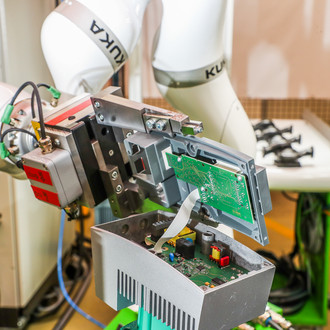 Das Bild zeigt eine Nahaufnahme eines industriellen Roboters, der an einer elektronischen Baugruppe arbeitet. Der Roboterarm ist weiß und trägt das Logo des Herstellers KUKA. Der Roboter hält eine teilweise geöffnete Baugruppe, die grüne Leiterplatten und verschiedene elektronische Komponenten enthält. Kabel und mechanische Teile sind ebenfalls sichtbar. Im Hintergrund sind weitere industrielle Ausrüstungen und Arbeitsbereiche zu sehen. Der Arbeitsplatz ist gut organisiert und deutet auf eine präzise und technische Umgebung hin.