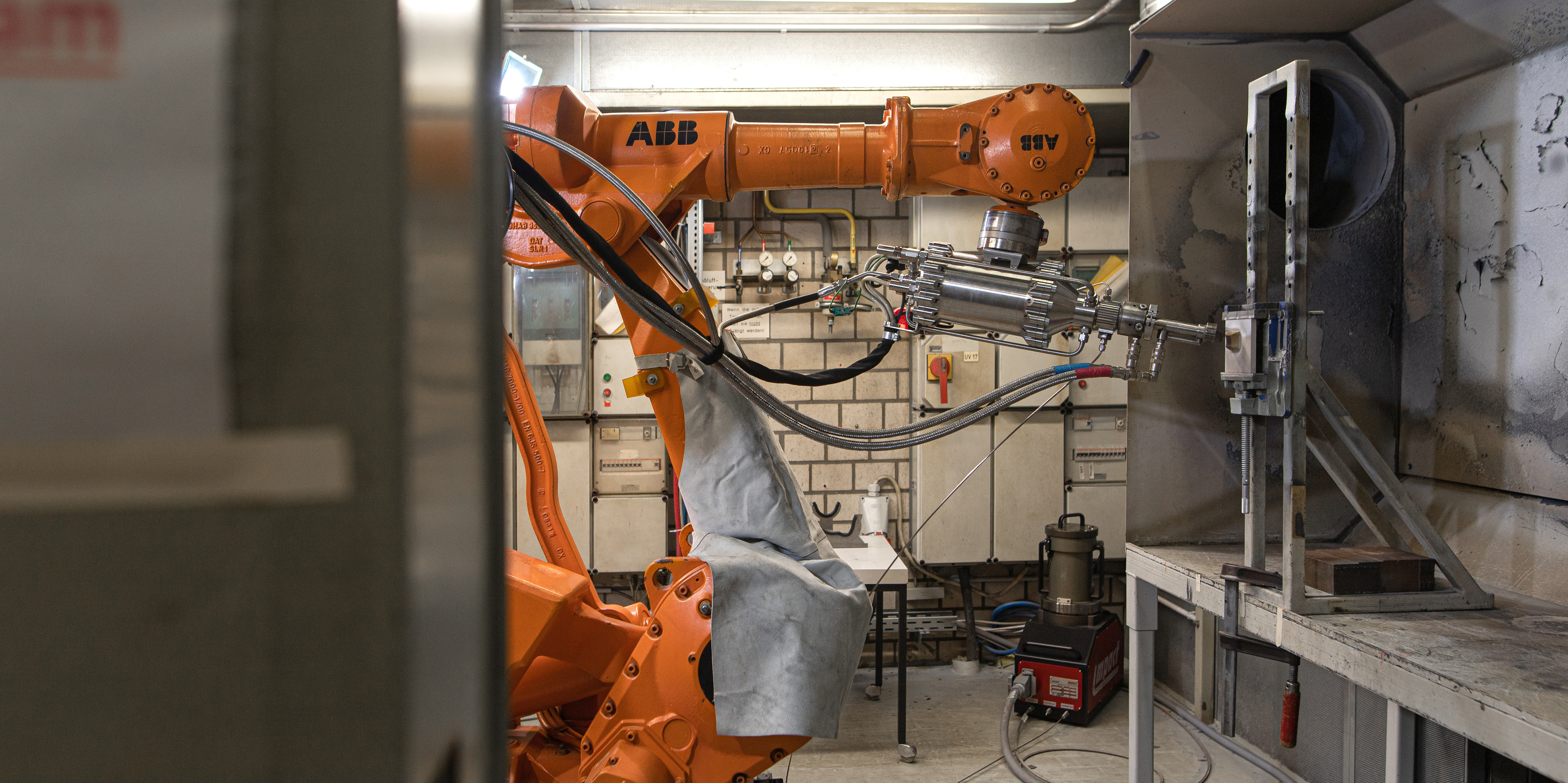 Foto eines industriellen Arbeitsbereichs mit einem orangefarbenen ABB-Roboterarm. Der Roboterarm ist in einem engen Raum positioniert und führt präzise Arbeiten an einer Vorrichtung durch, die an der rechten Wand montiert ist. Im Hintergrund sind Kabel, Werkzeuge und industrielle Ausrüstungen zu sehen. Der Raum wirkt funktional und ist durch helle Leuchtstofflampen beleuchtet. Der Boden ist staubig und der gesamte Bereich vermittelt den Eindruck einer aktiven Werkstattumgebung.