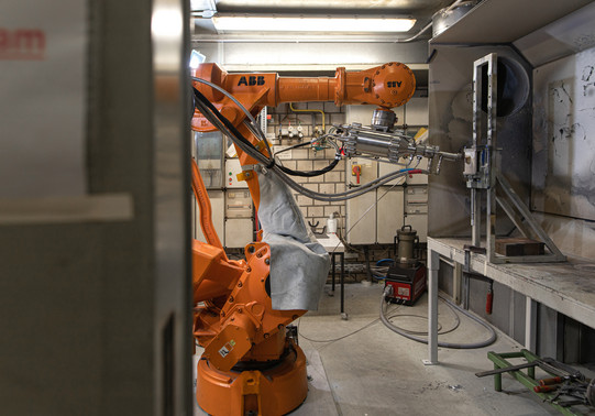 Foto eines industriellen Arbeitsbereichs mit einem orangefarbenen ABB-Roboterarm. Der Roboterarm ist in einem engen Raum positioniert und führt präzise Arbeiten an einer Vorrichtung durch, die an der rechten Wand montiert ist. Im Hintergrund sind Kabel, Werkzeuge und industrielle Ausrüstungen zu sehen. Der Raum wirkt funktional und ist durch helle Leuchtstofflampen beleuchtet. Der Boden ist staubig und der gesamte Bereich vermittelt den Eindruck einer aktiven Werkstattumgebung.