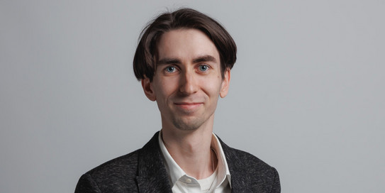 Das Bild zeigt ein Porträt eines jungen Mannes mit dunklem, kurzen Haar. Er trägt ein dunkelgraues Sakko über einem hellblauen Hemd und lächelt in die Kamera. Im Hintergrund sind große grüne Buchstaben „IPS“ zu sehen, die teilweise durch den Mann verdeckt werden. Der Hintergrund ist weiß und schlicht gehalten.