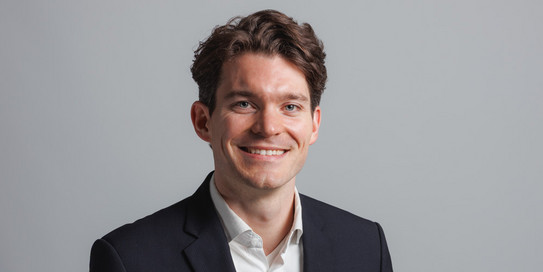 Ein Mann in einem grauen Anzug mit Krawatte lächelt in die Kamera. Er hat braune Haare und trägt einen Bart. Im Hintergrund sind große, grüne Buchstaben 'IPS' zu sehen, die teilweise durch den Mann verdeckt werden.