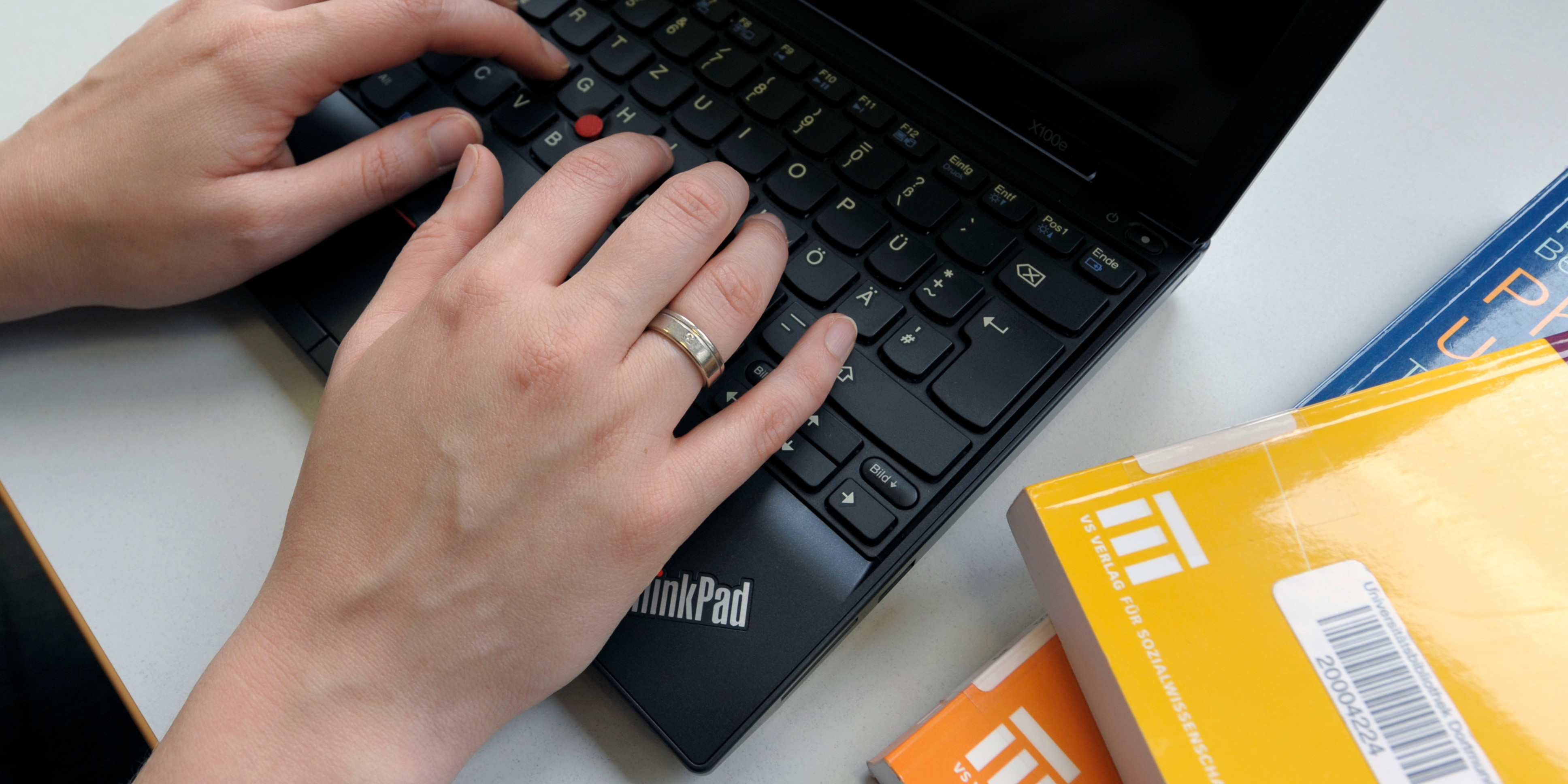 Das Bild zeigt die Hände einer Person, die auf einem ThinkPad-Laptop tippt. Die linke Hand trägt einen Ring am Ringfinger. Neben dem Laptop liegen mehrere Bücher, von denen zwei orangefarbene Einbände mit dem Logo eines Instituts oder einer Organisation haben. Der Arbeitsplatz wirkt sauber und organisiert.