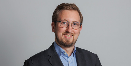Foto eines lächelnden Mannes im Anzug mit Krawatte, der vor einem Hintergrund mit den großen Buchstaben „IPS“ steht. Der Mann hat hellbraunes, nach hinten gekämmtes Haar und einen Bart. Die Buchstaben im Hintergrund sind in hellgrüner Farbe gehalten und heben sich deutlich vom weißen Hintergrund ab. Der Mann blickt direkt in die Kamera und strahlt Selbstbewusstsein aus.