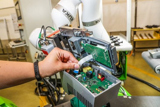 Foto eines Roboters, der bei der Montage eines elektronischen Geräts hilft. Eine menschliche Hand führt ein graues Kabel in eine grüne Leiterplatte ein, die sich in einer metallischen Halterung befindet. Im Hintergrund sind der Roboterarm und ein industrielles Umfeld zu sehen. Der Roboterarm ist weiß und trägt die Aufschrift "KUKA".
