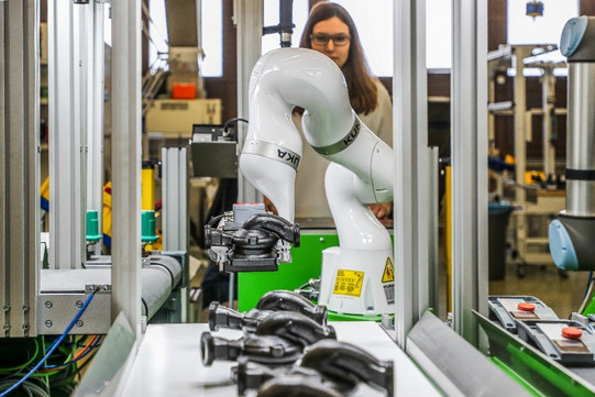 Das Bild zeigt eine industrielle Szene mit einem weißen KUKA-Roboterarm, der über einem Förderband arbeitet. Auf dem Förderband befinden sich mehrere schwarze mechanische Bauteile. Im Hintergrund ist eine Frau mit langen, braunen Haaren und Brille zu sehen, die den Roboterarm beobachtet. Die Umgebung ist eine moderne industrielle Einrichtung mit verschiedenen technischen Geräten und Maschinen.