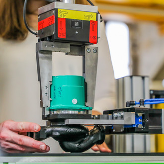 Das Bild zeigt eine Nahaufnahme einer industriellen Maschine, die ein mechanisches Bauteil bearbeitet. Die Maschine hält ein grünes Bauteil, während die Hände einer Person, die im Hintergrund zu sehen ist, die Maschine bedienen. Die Umgebung wirkt technisch und industriell mit unscharfen Hintergrundelementen.