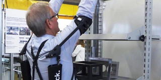 Foto eines Mannes in einem weißen Hemd, der in einer Werkstatt oder einem Labor an einem Regal arbeitet. Er trägt ein schwarzes Exoskelett an seinen Armen und Schultern, das ihn bei der Hebearbeit unterstützt. Der Mann hat graue Haare und trägt eine Brille. Im Hintergrund sind weitere Ausrüstungsgegenstände sowie Poster mit technischen Informationen zu sehen. Die Szene zeigt die Nutzung ergonomischer Hilfsmittel zur Unterstützung körperlicher Tätigkeiten in einer professionellen Umgebung.