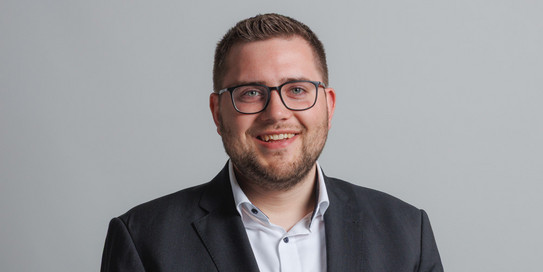 Das Bild zeigt einen jungen Mann mit kurzen, braunen Haaren und einer Brille, der in einem schwarzen Anzug mit weißem Hemd gekleidet ist. Er lächelt freundlich in die Kamera. Im Hintergrund ist ein großer, grüner Buchstabe "S" auf weißem Grund zu sehen, der Teil des Schriftzugs "IPS" ist.
