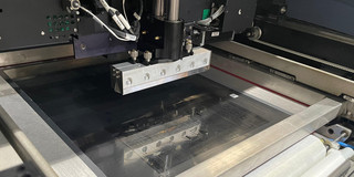Auf dem Foto ist ein industrieller Lotpastendrucker zu sehen, der gerade eine Leiterplatte (PCB) bearbeitet. In der Mitte befindet sich eine Metallrakel, die Lotpaste über eine Schablone streicht. Die Anlage ist präzise aufgebaut, mit sichtbaren Schrauben, Kabeln und mechanischen Komponenten, die den automatisierten Druckprozess unterstützen. Die Arbeitsfläche zeigt deutlich Spuren der aufgetragenen Lotpaste.