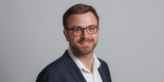 Foto eines jungen Mannes vor einem Hintergrund mit den Buchstaben "IPS". Der Mann hat kurzes, braunes Haar und trägt eine Brille mit schwarzem Rahmen. Er trägt ein hellblaues Hemd und ein dunkles Sakko und lächelt freundlich in die Kamera. Der Hintergrund ist in hellen Farben gehalten, wobei die Buchstaben "IPS" in einem großen, grünen Schriftzug zu sehen sind.