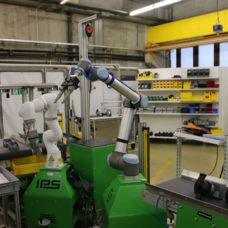 Innenaufnahme eines industriellen Labors oder einer Werkstatt mit zwei grünen, mobilen Roboterarmen, die verschiedene Aufgaben ausführen. Im Hintergrund sind Regale mit Werkzeugen und anderen Arbeitsmaterialien zu sehen. Die Roboter sind auf grünen Plattformen mit den Aufschriften 'IPS' und 'ISY' montiert. Die Umgebung ist gut beleuchtet mit industriellen Leuchten an der Decke.
