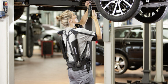 Foto einer Mechanikerin in einer Werkstatt, die an einem aufgebockten Auto arbeitet. Die Mechanikerin trägt ein graues Exoskelett, das ihre Bewegungen unterstützt. Sie hält ein Werkzeug in der Hand und arbeitet unter dem Fahrzeug, dessen Reifen und Unterboden gut sichtbar sind. Im Hintergrund sind weitere Fahrzeuge und Werkstattausrüstungen zu sehen. Die Szene zeigt eine moderne Arbeitsumgebung, in der ergonomische Hilfsmittel zur Unterstützung der körperlichen Arbeit verwendet werden.