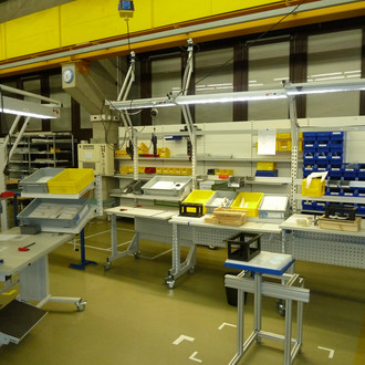Foto einer industriellen Werkstatt. Mehrere Arbeitsstationen mit weißen Tischen und Regalen sind entlang einer Wand aufgereiht. Die Arbeitsstationen sind mit gelben und blauen Behältern, Werkzeugen und Bauteilen ausgestattet. Über den Tischen hängen Leuchtstofflampen, und an den Stationen befinden sich elektrisch betriebene Werkzeuge. Der Boden ist hell und die Decke besteht aus gelben Strukturelementen. Im Hintergrund sind weitere Regale und industrielles Equipment zu sehen.