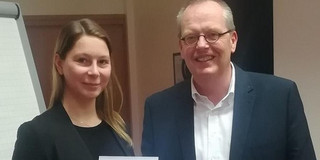 Foto eines Mannes und einer Frau in einem Büro. Die Frau hält ein Dokument in der Hand und trägt einen schwarzen Blazer, während der Mann in einem dunkelblauen Anzug und weißem Hemd gekleidet ist. Beide lächeln in die Kamera. Im Hintergrund ist ein Flipchart zu sehen. Die Szene wirkt formell und professionell, was auf eine geschäftliche oder akademische Veranstaltung hinweist.