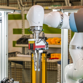 Foto eines weißen KUKA-Roboterarms in einer industriellen Umgebung. Der Roboterarm ist mit einem Greifer ausgestattet und befindet sich in der Nähe eines Arbeitsplatzes mit gelben Behältern im Hintergrund. Der Fokus liegt auf dem präzisen Greifmechanismus des Roboters, der auf einer modernen Produktionslinie eingesetzt wird. Die Szene ist gut beleuchtet und zeigt die fortschrittliche Technologie in einer industriellen Fertigungshalle.