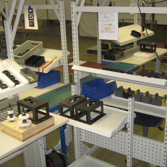 Foto: Ein gut organisierter Arbeitsbereich in einer Werkstatt mit mehreren Regalen und Arbeitsflächen. Auf den Tischen und Regalen befinden sich verschiedene Werkzeuge, Bauteile und Materialien, die ordentlich sortiert und platziert sind. Auf einem Tisch liegt eine Holzplattform mit montierten Bauteilen, daneben stehen Metallrahmen. Im Hintergrund sind weitere Arbeitsbereiche und Tische zu sehen, die mit ähnlichen Materialien bestückt sind. Die Umgebung wirkt sauber und funktional, ideal für präzise Montagearbeiten.
