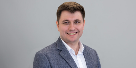 Foto eines jungen Mannes vor einem Hintergrund mit den Buchstaben "IPS". Der Mann hat kurzes, braunes Haar und trägt ein weißes Hemd sowie ein graues Sakko. Er lächelt freundlich in die Kamera. Der Hintergrund ist in hellen Farben gehalten, wobei die Buchstaben "IPS" in einem großen, grünen Schriftzug zu sehen sind.