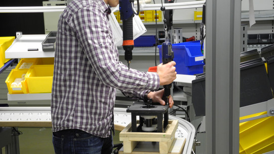 Foto einer Person, die in einer industriellen Umgebung an einem Förderband arbeitet. Die Person trägt ein kariertes Hemd und nutzt ein elektrisches Werkzeug, um ein Bauteil auf einem Holzbock zu befestigen. Im Hintergrund sind Regale mit gelben und blauen Behältern zu sehen, die verschiedene Materialien und Werkzeuge enthalten. Die Person ist von der Seite zu sehen und ihr Gesicht ist nicht erkennbar.