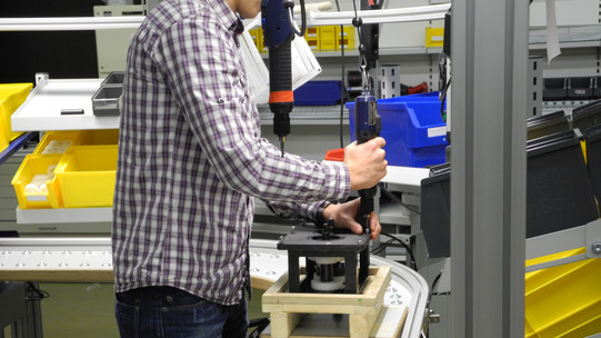 Foto einer Person, die in einer industriellen Umgebung an einem Förderband arbeitet. Die Person trägt ein kariertes Hemd und nutzt ein elektrisches Werkzeug, um ein Bauteil auf einem Holzbock zu befestigen. Im Hintergrund sind Regale mit gelben und blauen Behältern zu sehen, die verschiedene Materialien und Werkzeuge enthalten. Die Person ist von der Seite zu sehen und ihr Gesicht ist nicht erkennbar.