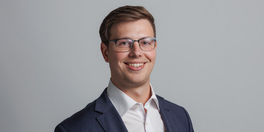 Porträtfoto eines lächelnden jungen Mannes mit kurzen, seitlich gestylten braunen Haaren, der ein dunkelgraues Sakko, ein hellblaues Hemd mit Punkten und eine dunkelblaue Krawatte trägt. Der Hintergrund zeigt die Buchstaben 'IPS' in großen, grünen und grauen Blockbuchstaben.