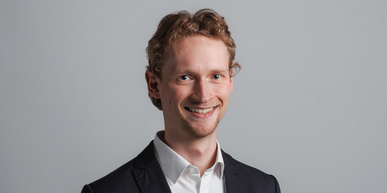 Foto eines lächelnden Mannes in einem schwarzen Anzug mit weißem Hemd. Der Mann hat kurze, wellige Haare und blickt direkt in die Kamera. Im Hintergrund ist ein großes, teilweise verdecktes Logo mit den grünen Buchstaben "IPS" auf weißem Hintergrund zu sehen.