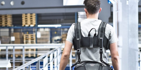 Das Bild zeigt einen Mann von hinten, der ein Exoskelett trägt. Der Mann trägt ein graues T-Shirt und befindet sich in einer industriellen Umgebung mit Regalen und Paletten im Hintergrund. Das Exoskelett ist schwarz und unterstützt den Rücken und die Schultern des Mannes.