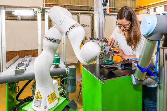 Foto einer Frau, die in einer industriellen Umgebung an einer Maschine arbeitet. Die Frau trägt eine Brille und konzentriert sich auf ein Bauteil, das sie bearbeitet. Neben ihr befinden sich zwei Roboterarme, einer davon mit dem Markennamen KUKA. Die Roboterarme sind an einer grünen Arbeitsstation befestigt. Der Hintergrund zeigt weitere industrielle Ausrüstung und eine helle, gut beleuchtete Werkstattumgebung.