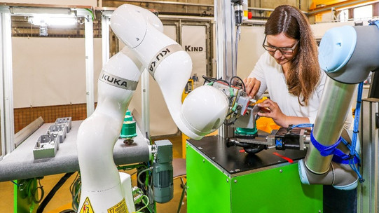 Foto einer Frau, die in einer industriellen Umgebung an einer Maschine arbeitet. Die Frau trägt eine Brille und konzentriert sich auf ein Bauteil, das sie bearbeitet. Neben ihr befinden sich zwei Roboterarme, einer davon mit dem Markennamen KUKA. Die Roboterarme sind an einer grünen Arbeitsstation befestigt. Der Hintergrund zeigt weitere industrielle Ausrüstung und eine helle, gut beleuchtete Werkstattumgebung.