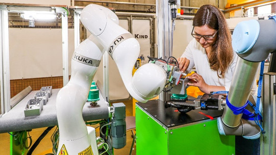 Foto einer Frau, die in einer industriellen Umgebung an einer Maschine arbeitet. Die Frau trägt eine Brille und konzentriert sich auf ein Bauteil, das sie bearbeitet. Neben ihr befinden sich zwei Roboterarme, einer davon mit dem Markennamen KUKA. Die Roboterarme sind an einer grünen Arbeitsstation befestigt. Der Hintergrund zeigt weitere industrielle Ausrüstung und eine helle, gut beleuchtete Werkstattumgebung.