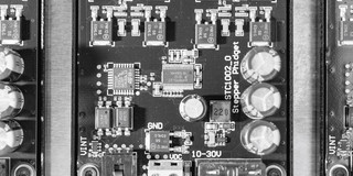 Foto einer Leiterplatte mit mehreren elektronischen Bauteilen, darunter Kondensatoren, Widerstände, integrierte Schaltungen und Schraubklemmen. Die Platine ist symmetrisch aufgebaut, in der Mitte befindet sich ein größerer Chip, umgeben von weiteren Komponenten, die in strukturierter Anordnung verlötet sind. Rechts sind drei größere Elektrolytkondensatoren sichtbar, während oben mehrere blaue Steckbrücken und kleinere Bauteile angeordnet sind.