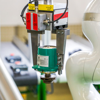 Foto eines Roboters, der ein industrielles Bauteil hält. Der Roboterarm ist weiß und trägt die Aufschrift "KUKA". Er hält mit einer Greifvorrichtung ein grünes, zylindrisches Bauteil, das detailliert beschriftet ist. Im Hintergrund sind unscharfe industrielle Geräte und Maschinen zu sehen, die eine Werkstatt oder Produktionsumgebung andeuten.