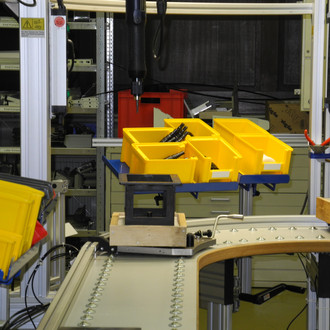 Foto eines Arbeitsplatzes in einer industriellen Produktionsumgebung. Im Vordergrund verläuft ein Förderband, das um eine Kurve führt. Auf dem Förderband steht ein Halter mit mehreren gelben Kunststoffbehältern, die verschiedene Werkzeuge und Materialien enthalten. Links im Bild befinden sich weitere gelbe Behälter, die auf einem Regal gestapelt sind. Im Hintergrund sind verschiedene Maschinen, Regale und ein roter Behälter zu sehen. Der Arbeitsplatz ist von einem Aluminiumrahmen mit integrierter Beleuchtung umgeben, der eine strukturierte und gut organisierte Arbeitsumgebung schafft.